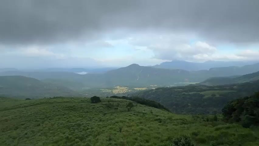 The video shows a wide, green, grassy hill with sparse trees in the foreground. In the distance, there are several hills and mountains, fading into the background with a body of water visible between them. The sky is overcast with grey and white clouds, creating a dramatic atmosphere. There are no people visible in the scene. The camera appears to be stationary, providing a scenic overview of the natural landscape.