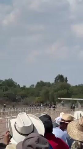 The video shows a horse race with a crowd watching, mostly men wearing hats, and some taking photos. The race is happening on a dirt track with a white metal barrier and trees in the background. It's a sunny day with a partly cloudy sky. The horses are kicking up dust as they race.