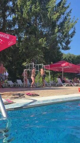 The video shows an outdoor area with a swimming pool in the foreground. In the middle ground, two people are standing under outdoor showers. A woman in a black bikini is facing away from the camera, and a man in black swim trunks is facing her. Behind them, there are lounge chairs, trees, and red umbrellas with Coca-Cola logos. The man is speaking to the woman. The scene appears to be a sunny day.