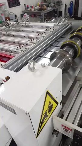 The video shows a printing machine in operation. The machine has a roll of red material with white text and logos being fed through it. A large metal roller with yellow rings is seen to be cutting the material. The machine appears to be in a workshop or factory setting, with tools and other equipment visible in the background. There are also warning labels on the machine, indicating potential hazards. The video is taken from a close-up perspective, focusing on the printing and cutting mechanism.