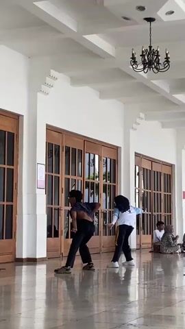 Two people, a male and a female, are dancing on a covered walkway in front of a large building with wooden doors. The male is wearing a dark-colored shirt, but the shirt is pulled up revealing his back and stomach, dark pants, and shoes. The female is wearing a white jacket with light blue accents, dark pants with yellow stripes, white shoes, and a dark head covering. Both are in motion, facing the doors. Behind them and to the right, several individuals sit on the floor. Above them is a white ceiling with decorative elements and a black chandelier. The floor is a shiny, polished surface. A red and white sign is posted on the doors to the left of the dancers.