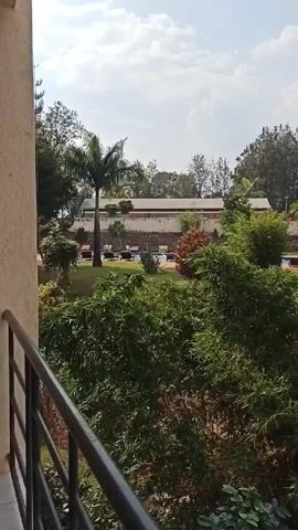 The camera pans right across a verdant outdoor scene from an elevated position, likely a balcony.  In the foreground, the gray balcony railing is visible with dense green bushes and trees filling the lower part of the frame. Beyond the foliage lies a well-manicured lawn. A swimming pool with lounge chairs around it is partially visible in the center. Further back, a building with a light-colored facade and brown accents sits in the distance. To the right, a gazebo-like structure with a blue roof is prominent, connected to another blue-roofed walkway.  Various trees and plants are scattered throughout the area. The sky is a light blue with some white clouds. Sounds of music can be heard in the background.