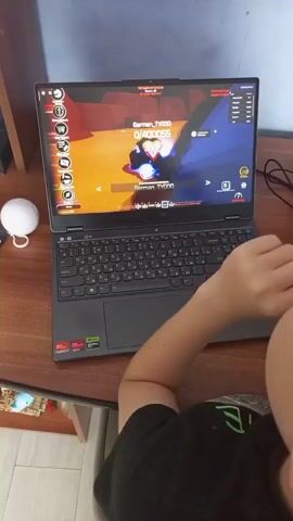 The video captures a person playing a video game on a laptop. The laptop is open and placed on a wooden desk. The game displayed on the screen appears to be an action-oriented game with vibrant graphics, featuring characters in combat and bright visual effects. The person's right arm and hand are visible, with their fingers positioned near the keyboard and trackpad, indicating active gameplay. To the left of the laptop, a small white smart speaker with a light ring is present. The background is slightly out of focus but suggests an indoor setting, possibly a room with a wall and some shelving. The overall scene is typical of someone engaged in a gaming session.