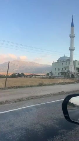 This media file is a vertical video showcasing a serene landscape with a mosque as a prominent feature. The scene transitions from a view of the mosque and its minaret, with mountains in the background, to a broader view of a rural or semi-rural area. The time of day appears to be late afternoon or early evening, indicated by the soft, warm light and the long shadows. The sky is a clear blue with some wispy clouds, suggesting pleasant weather.

The mosque itself is a light green building with a tall, slender minaret topped with a blue spire. The architecture has traditional Islamic elements, including a dome and arched windows. The surrounding area is dry and somewhat dusty, with patches of tall, dry grass and some scattered trees. Power lines and utility poles are visible, suggesting the presence of infrastructure.

As the video progresses, the view expands to reveal more of the surroundings. We see a dirt road or track leading away from the main paved road in the foreground. In the distance, there are more trees and the faint outline of buildings. The mountains in the background are rugged and appear to be bathed in the warm light of the setting sun.

Later in the video, the scene shifts to show parked vehicles, including trucks and a smaller car, in what looks like a lot or a service area. There is also a piece of construction equipment, possibly an excavator, visible. This suggests a mix of residential, religious, and possibly industrial or agricultural activity in the area.

There is no visible text in the media file. The overall mood is peaceful and calm, evoking a sense of rural tranquility. The location cues, such as the mosque and the mountainous backdrop, combined with the time of day and the general aesthetic, point towards a setting in a region like Seydikemer, Turkey.
