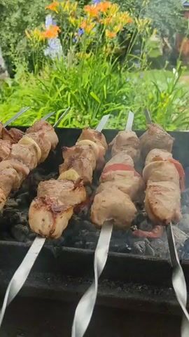 This video shows skewers of meat being grilled over coals. The meat appears to be pork and is interspersed with pieces of onion and possibly tomato. Smoke rises from the grill, indicating that the meat is cooking. In the background, there are green plants and some orange and yellow flowers. The overall scene suggests an outdoor barbecue or picnic.