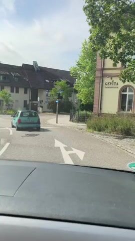 The video shows a view from the inside of a car, looking forward through the windshield. The car is stopped at an intersection with a traffic light. A light green car is in front of the camera car, also stopped. There are other cars on the road, and buildings are visible on either side of the street. The sky is partly cloudy. The traffic light is red.