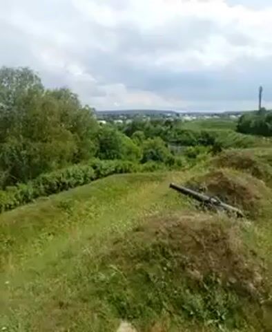 The video shows a panoramic view of a historical site with cannons. The camera pans from left to right, revealing the landscape. In the foreground, there are grassy mounds, with a cannon visible on one of them. Further in the distance, there are trees, buildings, and a cloudy sky. The overall scene suggests an outdoor historical or archaeological location, possibly a fortification or a museum exhibit. The video appears to be filmed on a handheld device, with occasional slight movements. There are no people visible in the video, and the focus is on the environment and the historical artifacts.