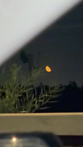 A bright orange, crescent-shaped object hovers in a dark sky, appearing slightly out of focus. In the foreground, blurred green and dark shapes suggest vegetation or a fence line. The object's glow illuminates the surrounding darkness.