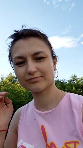 This media file features a young woman with dark hair, wearing a pink tank top with text on it, and hoop earrings. She is outdoors, in what appears to be a rural setting. The background consists of lush green trees and foliage, a dirt road, and a cloudy sky with hints of blue. There are also utility poles visible in the distance.

The woman's expression is neutral, perhaps a slight smile. Her hair is pulled back loosely, and she is looking directly at the camera. The overall atmosphere suggests a casual, daytime activity. The text on her shirt appears to be in a stylized font, with some letters in white and a prominent pink script underneath.

The scene is likely Tashkazgan, Russia, given the context. The time of day seems to be late afternoon or early evening, judging by the lighting and sky. The weather appears to be mild, with clouds suggesting it might be partly sunny. The dirt road implies a less developed area, possibly a village or countryside.