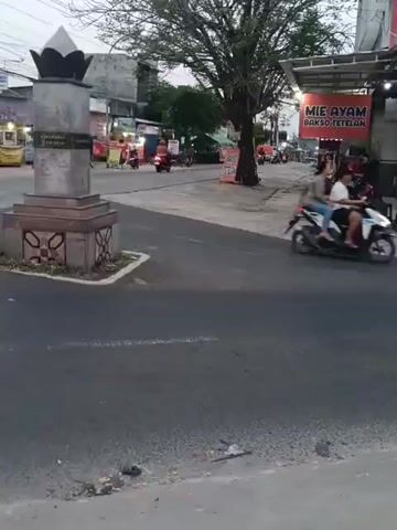 This video captures a street scene, likely in a populated area, with a roundabout in the foreground. A monument stands in the center of the roundabout. The road surface is asphalt, with some debris visible at the bottom of the frame. On the right side of the road, there is a building with a sign that reads "MIE AYAM BAKSO TETELAN," indicating a food establishment. Several motorcycles are present, with some individuals riding them, suggesting traffic activity. The background shows buildings and trees, typical of an urban or suburban environment. The lighting suggests it might be late afternoon or early evening.