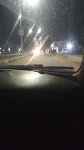 A first-person perspective video captures a nighttime drive through a dimly lit urban area. The camera is positioned inside a vehicle, showing the dashboard and windshield. The windshield is dirty, with streaks and spots visible, and appears to have water or rain on it, creating light flares from streetlights and oncoming headlights. The road ahead is visible, illuminated by streetlights and the headlights of other vehicles. Several streetlights are visible on poles along the side of the road. There are some structures and buildings on the right side of the road. In the distance, there are a few figures walking or standing on the side of the road, and some vehicles can be seen. The overall atmosphere is dark and slightly hazy.