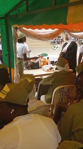This video shows a group of people under a green and yellow tent. In the foreground, several individuals are seated, facing away from the camera, wearing green uniforms and hats. A young woman in a white t-shirt with "NYSC" on it and green pants stands at a table, handing an item to a woman in a black abaya and hijab. The table is laden with various items, including bottles, containers, and a dark bag. In the background, more people are visible, some in similar uniforms, and beyond them, there appears to be an outdoor gathering with chairs and a white structure adorned with flowers. The setting suggests an organized event, possibly a ceremony or distribution under a temporary canopy. The lighting indicates it is daytime, and the atmosphere seems formal. Visible text on a chair in the foreground reads "N.Y.S.C." and "COURTLY ENAMS."