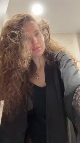 A woman is blow-drying her hair in what appears to be a bathroom. She's wearing a black top and a dark robe with lace sleeves. She's holding the hair dryer with her left hand and uses her right to lift and fluff her long, curly hair. The background shows tiled walls, a towel rack, and a ceiling light.