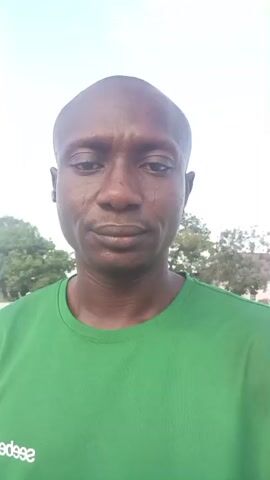 The video shows a close-up, head-and-shoulders shot of a dark-skinned man wearing a green t-shirt. He has a shaved head and appears to be outside, with trees and buildings visible in the background. The man's expression is neutral.
