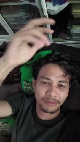 The video shows a man with dark hair and a mustache looking at the camera. He is wearing a dark t-shirt. His left hand is raised to his head, touching his hair. The background appears to be a room with shelves filled with books and papers, and a ceiling fan. The lighting is dim.