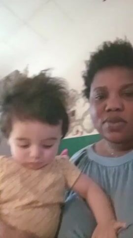 A woman holds a young child in a home. The woman speaks into the camera, while the child looks down with a somber expression. The child wears a brown shirt with a diamond pattern. The woman wears a light-blue shirt with a gathered neckline. They sit in front of a wall with floral wallpaper, and a white tiled ceiling is visible overhead.