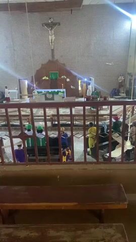 The video shows an interior view of a church, looking towards the altar. In the center of the frame, a large crucifix depicting Jesus Christ is mounted on the wall above the altar. The altar itself is adorned with white and green cloths, and lit candles. Several people are visible in the church. In the foreground, there are wooden benches. Behind a railing, a group of people are seated on benches, facing the altar. Some are wearing green head coverings. Towards the altar, several individuals in clerical attire appear to be conducting a service. The walls of the church are tiled, and there are large windows or openings allowing light to enter, creating beams of light visible in the frame.