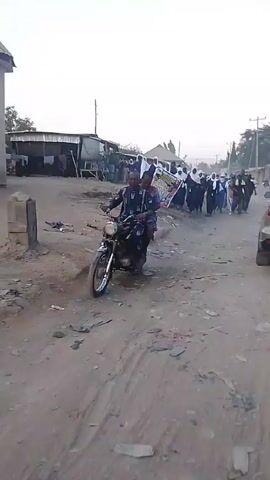 The video shows a group of people walking down a dirt road. Most of the women are wearing white hijabs and dark blue or black dresses, with a few men in regular clothing mixed in. They are carrying a sign that appears to be a memorial or tribute. The road is dirty and unpaved with bits of trash strewn about. In the background, there are simple buildings and a car parked on the side of the road. A man wearing a baseball cap, vest and carrying a camera bag runs past the group. The overall setting suggests a low-income urban or semi-urban area. The lighting indicates that it is daytime, likely in the morning or late afternoon.