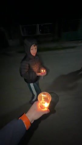 Here is a description of the video:
The video shows two people holding candles outside at night. The person holding the camera has an orange smartwatch on their wrist and is holding a lit candle in a clear plastic cup. A child in a black hooded jacket is walking away, holding a lit candle in their hands. The area is dark, with the candles providing the only light. There's a building with windows visible in the background. The child turns to look at the camera.