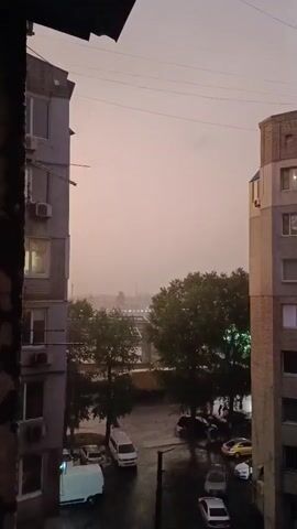 This is a short, vertical video showing an apartment building courtyard during a rainstorm. The sky is overcast and a hazy, orange light is visible in the distance. It is raining heavily, and the pavement is wet and reflects the light from streetlamps. There are several cars parked in the courtyard, including a white van. Trees are visible in the background, and some have green lights shining on them. The video is filmed from a window in another apartment building, with the edge of the building visible in the foreground. The audio features the sound of heavy rain and distant thunder.