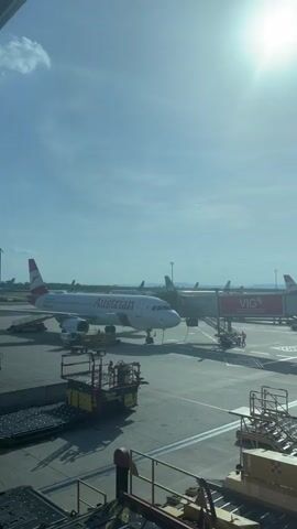 The video shows a view from inside an airport terminal, looking out onto the tarmac on a bright, sunny day. Visible are several airplanes, including an Austrian Airlines plane at a gate labeled VIG. Various airport vehicles and equipment are scattered across the tarmac. The camera pans slightly to the right, revealing an elevated roadway used for airport traffic, with yellow service vehicles and trucks moving along it. The building architecture is modern, with glass and metal structures. No people are distinctly visible in the video beyond ground staff around the planes.