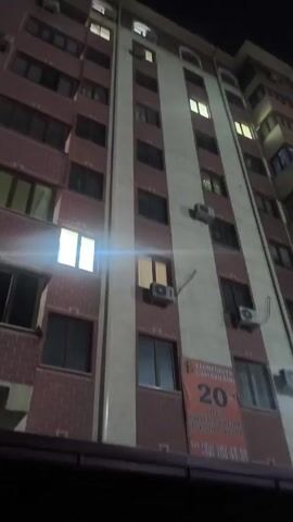 The video depicts the exterior of a tall building at night. The building has multiple floors, with rows of windows, some of which are lit. Air conditioning units are visible on the exterior walls of the building. A sign with the word "FLORENSIYA" is present. The camera pans upwards, capturing the dark sky above with a few faint stars and some wispy clouds. Bright streaks of light cross the sky, possibly from a light source or reflection.