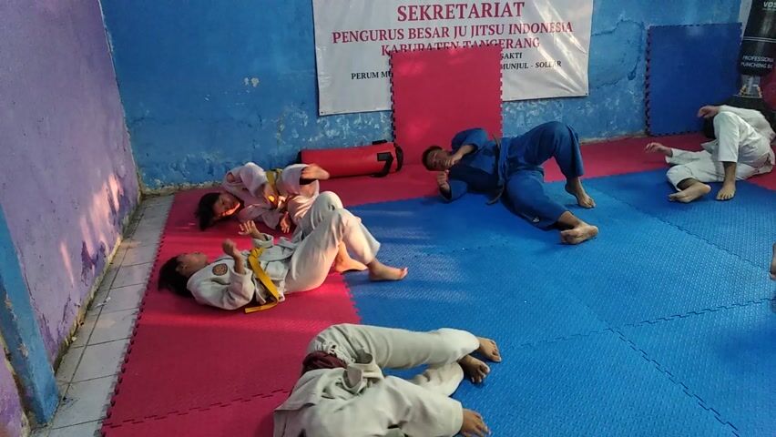 The video depicts a group of people, likely children and young adults, practicing martial arts on a blue and red mat-covered floor. They are wearing martial arts uniforms, known as gis, in various colors like white, blue, and black. The individuals are in different poses, some lying down, some sitting, and one person in a blue gi appears to be in motion. The scene is indoors, in what appears to be a dojo or training facility. The walls are painted blue and red, and there are windows with an outdoor view of trees and what looks like a residential area. A large banner with Indonesian text is visible on the back wall, which reads "SEKRETARIAT PENGURUS BESAR JIJIU JITSU INDONESIA KABUPATEN TANGERANG." There are also training equipment like a punching bag and gym bags present in the room. The overall atmosphere seems to be one of training and physical activity. The lighting suggests it could be daytime, though there are no clear indicators of the time of day or weather conditions. The focus is on the martial arts practice and the participants.