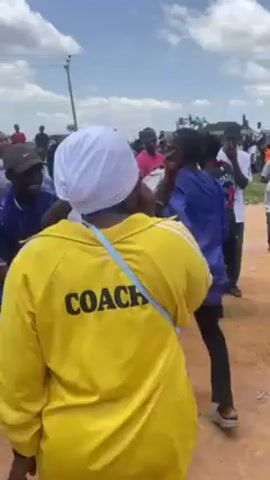 The video captures a lively outdoor scene filled with people dancing and celebrating. The environment appears to be a dirt road or open area under a partly cloudy sky. There are numerous people of various ages present, most of whom are moving and dancing to music. Many of the people are wearing casual clothing, including athletic shirts and shorts. A woman in a yellow tracksuit with the word "COACH" printed on the back is prominent in the foreground. One individual is wearing a Chelsea football shirt. There's trash and scattered objects on the ground, suggesting this is an informal gathering. The overall atmosphere is festive and energetic.