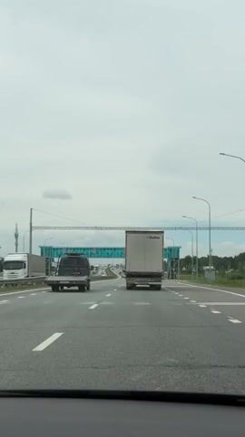 A first-person view from inside a car driving on a multi-lane highway on an overcast day. Ahead, there are two trucks, one in each lane, with a pedestrian bridge crossing above the road in the distance. The road is clear of other traffic besides the two trucks. The car is driving in the middle lane.