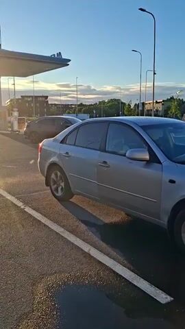 A silver sedan is parked in a parking lot. The car is viewed from the side, with the front of the car facing to the right. The car is in focus, and the background is slightly blurred. The sun is shining, casting a shadow of the car on the asphalt. In the background, there is another car with the Bolt logo on it. There are also some trees and buildings in the distance.