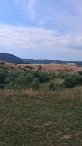 A drone footage captures a panoramic view of a rolling landscape under a partially cloudy sky. The camera pans across rolling hills covered in dry grass and scattered trees, leading to a cluster of denser green trees in the mid-ground. Beyond this, the terrain becomes more rugged, with hills and valleys, and a distant mountain range with a hazy blue hue. In the lower right corner, a group of people are seen walking along the grassy bank of a body of water. The camera then shifts focus to a close-up of the water's surface, showing ripples and reflections. The drone continues its flight, showcasing a vast expanse of water bordered by a dense forest that climbs up a steep hill. The sky overhead is a mix of blue and grey clouds.