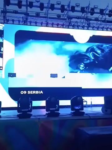 The video shows a large screen displaying a video of a man in what appears to be a music video. The screen is set up on a stage with stage lighting arranged in front. The lower left corner of the screen displays the text "09 SERBIA." The video on the screen features fast-paced visuals and appears to be accompanied by music.