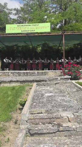A medium full shot captures a group of men and women performing a traditional dance on a stage under a green banner with Bulgarian text. They are dressed in white shirts, black vests, and red skirts or pants with embroidery. The stage is made of light-colored stone, and beyond it, there's lush green foliage and red flowers. In the foreground, there are stone steps leading down to a grassy area.