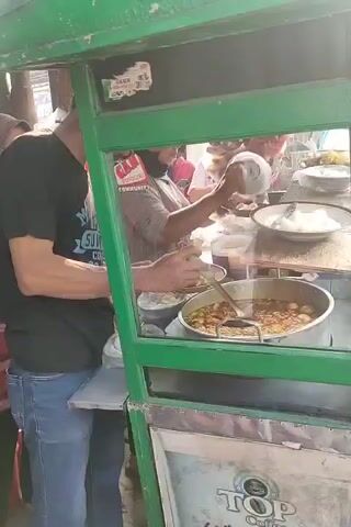 A vendor is preparing food at a street cart. The cart has a green frame and glass windows. In the foreground, a pot of stew with meatballs is visible, along with a bowl of white food items, possibly rice cakes or dumplings. The vendor, wearing a grey shirt and a black face mask, is shown scooping food into a bowl and adding something from a small green bottle. Another person is visible in the background, also interacting with the food. The scene appears to be outdoors, with bright sunlight suggesting daytime.