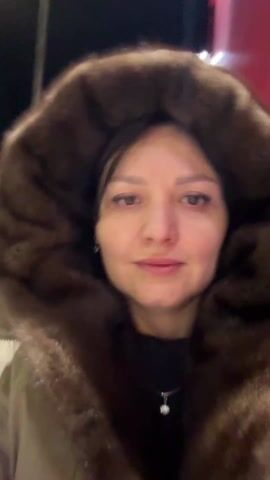 A woman wearing a brown fur-lined hood is walking outside at night. There is snow on the ground and streetlights illuminate the scene. The background includes cars and buildings. The woman appears to be looking directly at the camera. The video is a close-up shot.