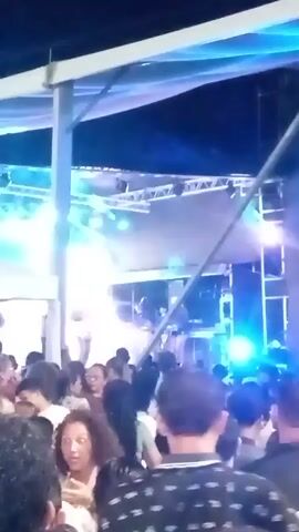 The video captures a lively outdoor concert in San Juan Bautista, Venezuela. A band is performing on a stage illuminated by vibrant blue and white lights. The lead singer, dressed in dark clothing and a cap, is playing a guitar. Other band members are visible behind him, along with musical equipment like drums.

A large crowd of people is gathered in front of the stage, enjoying the performance. Many are looking towards the stage, and some appear to be dancing or reacting to the music. The atmosphere seems energetic and celebratory. The concert is taking place at night, indicated by the darkness of the sky and the prominent stage lighting. The setting is partially covered by a roof structure, suggesting it's an open-air venue. The crowd is diverse, with people of various ages and genders. The overall impression is of a fun and engaging musical event.