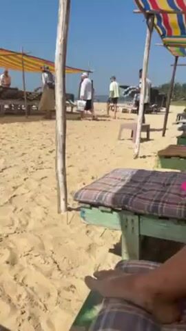 The video shows a sandy beach on a bright, sunny day. In the foreground, there is a wooden seating area with a cushion. The camera is positioned from the perspective of someone sitting down, showing a person's foot on the edge of the seat. Beyond the seating area, the beach extends towards the ocean. There are several wooden structures with colorful striped canopies, possibly beach bars or shaded seating areas. People are scattered around the beach, some walking, others sitting on the wooden structures.