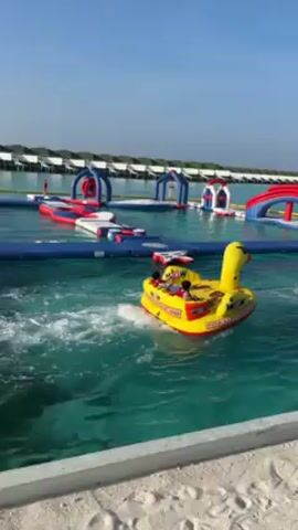 A person sits in the front of a yellow and red inflatable duck boat and rides in the water with another person in the back. The people move from left to right. In the background is an inflatable obstacle course on the water. Further back are small cabin-like structures on stilts. The sky is blue.