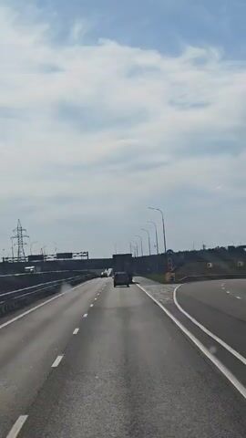 The video shows a view from a vehicle driving on a multi-lane highway. The highway has a concrete barrier separating lanes and a wide shoulder. There are several other vehicles on the road, including cars and trucks. The sky is partly cloudy, and there are streetlights and power lines visible along the side of the road. Road signs indicating lane closures and directional information are also present. The overall scene depicts typical highway traffic and infrastructure under a cloudy sky.