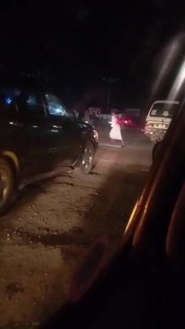 A dark-colored car with its brake lights illuminated is stopped on a dirt road at night. The car's rear tire appears to be partially deflated or damaged. Several other vehicles are visible in the background, and streetlights illuminate the scene. A person is standing next to the driver's side door of the dark-colored car, wearing patterned pants and a dark top. The camera is positioned inside another vehicle, looking out at the scene.