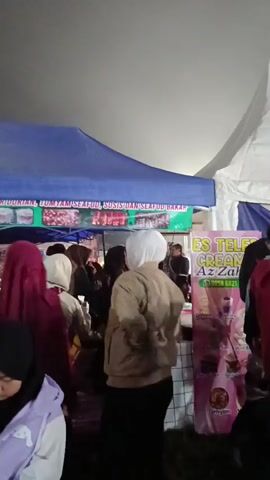 A night scene shows a bustling outdoor market with multiple tents. A blue tent in the background displays food items like seafood and sausages. A white tent is to the right. In the foreground, people of various ages are visible. A child in a green shirt and black jacket stands facing left, looking towards a stand. Another person wearing a light brown jacket and white head covering is also facing left. A purple tent is partially visible on the far left, with a person wearing a purple hoodie. A pink banner for "ES TELE CREAM Az-Zahra" is displayed on a stand, featuring images of drinks. The ground appears to be covered in dirt or debris. The lighting suggests it is nighttime, with artificial lights illuminating the area.