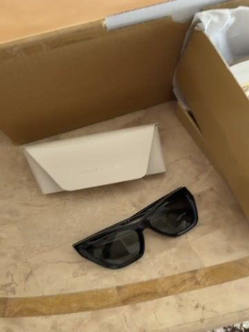 The video shows a collection of Michael Kors branded items on a light wooden surface. An open Michael Kors shoebox reveals a pair of white sneakers with beige accents and the MK logo. A brown Michael Kors shopping bag is placed inside a larger, white Michael Kors box. Beside the shoebox lies a white sunglasses case and a pair of black sunglasses. The backdrop includes a beige bedspread and pillows, suggesting the scene is indoors.