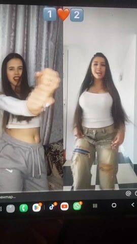Two young women are dancing in a split-screen video. The woman on the left wears a white long-sleeved crop top and gray sweatpants. She has long brown hair and stands in front of a draped curtain. The woman on the right wears a white tank top and ripped light-wash jeans. She also has long brown hair and stands in front of a white wall with a black and white tiled floor. Both women smile and make hand gestures as they dance. The video has overlaid text at the top indicating a voting format with the numbers 1 and 2, separated by a heart emoji. At the bottom, there are navigation icons and text in a foreign language. The video is displayed on a tablet device.