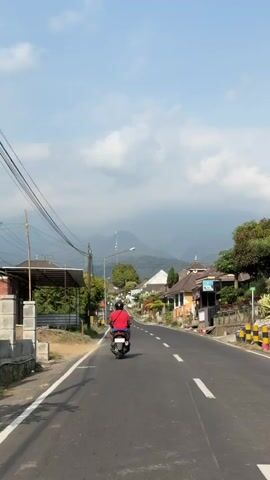 The media shows a point-of-view shot of someone riding a motorcycle down a paved road in Gubukklakah, Indonesia. The rider, wearing a red shirt and blue shorts, is in the foreground. The road is lined with buildings, some of which appear to be homes with traditional architecture, and lush greenery. In the distance, mountains are visible, shrouded in mist, with a tall telecommunications tower standing out against the sky. The sky is a mix of blue and white clouds, suggesting it is daytime, likely late morning or early afternoon given the bright sunlight. The overall atmosphere appears peaceful and rural, with the rider moving through the village. There are no visible signs with text.