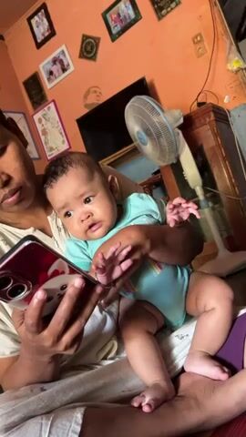 A close-up shot focuses on a baby being held by an adult. The baby, wearing a teal onesie with colorful stripes, looks directly at a smartphone held by the adult. The adult's hands, with visible fingernails, grip the phone. The phone's screen displays a cartoon character. The background shows a room with an orange wall, a fan, and framed pictures. The lighting is warm and natural. The baby's expression shifts from mild curiosity to a wide smile.
