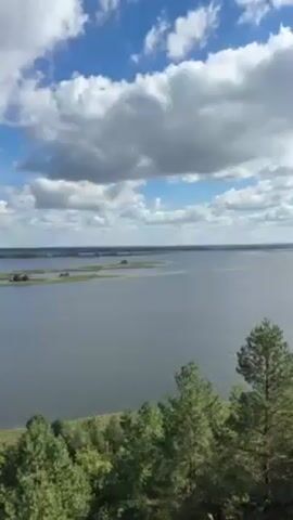The video shows a wide view of a large body of water under a partly cloudy blue sky. There are a few small islands visible in the distance. The foreground shows trees and foliage. The camera then shifts to a woman wearing sunglasses and a plaid shirt, taking a selfie with the lake in the background. She touches her hair.
