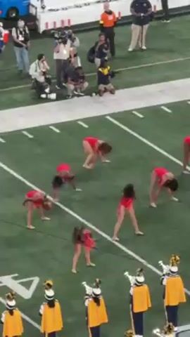 The video shows a group of dancers in red leotards performing on a green football field. They are performing in front of a marching band in blue and gold uniforms. There are various people in the background, including photographers, security personnel, and spectators. The dance formation shifts as the music plays, at one point with the dancers performing synchronized rolls. The scene appears to be part of a halftime show or pre-game performance at a sporting event.