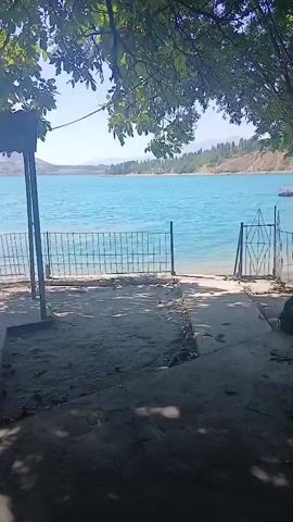 A video shows a body of turquoise water with a fence in the foreground and trees overhead. The camera pans slowly to the right. There is a path leading to the water, and a gate in the fence. The water appears calm and there are some trees and hills in the background.