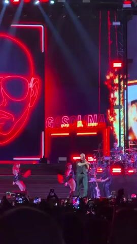 This is a video of a live concert performance. A male singer is on stage, wearing a dark outfit and sunglasses. He is holding a microphone and appears to be singing. There are two female dancers on stage with him, wearing skimpy outfits. Behind the singer, there is a large screen displaying a neon outline of a man's face wearing sunglasses and the word "GASOLINA" in neon letters. The stage is lit with red and blue lights. There is a drum set and other musical equipment on stage. A crowd of people is visible in the foreground, holding up their phones to record the performance.