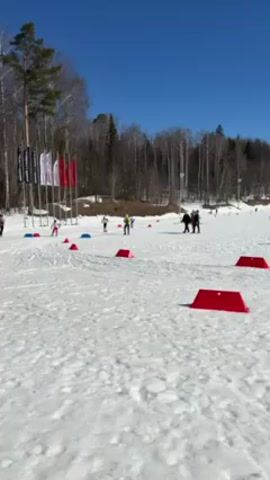 The video shows a cross-country skiing race on a sunny day. The ground is covered in snow and marked with red rectangular obstacles. Several skiers, wearing racing attire, are seen maneuvering through the course. In the background, there are trees and a blue sky. There are also several people standing around the course, likely watching the race. Some flags are also visible near the starting point of the course.